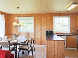 A wooden-paneled room with a dining table, chairs, a kitchen area with a microwave, and a window providing a view of greenery.