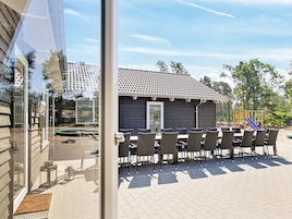 Un espace extérieur aménagé en terrasse avec une table et des chaises, un petit bâtiment doté d’un toit en tuiles, et un terrain de jeux en arrière-plan.