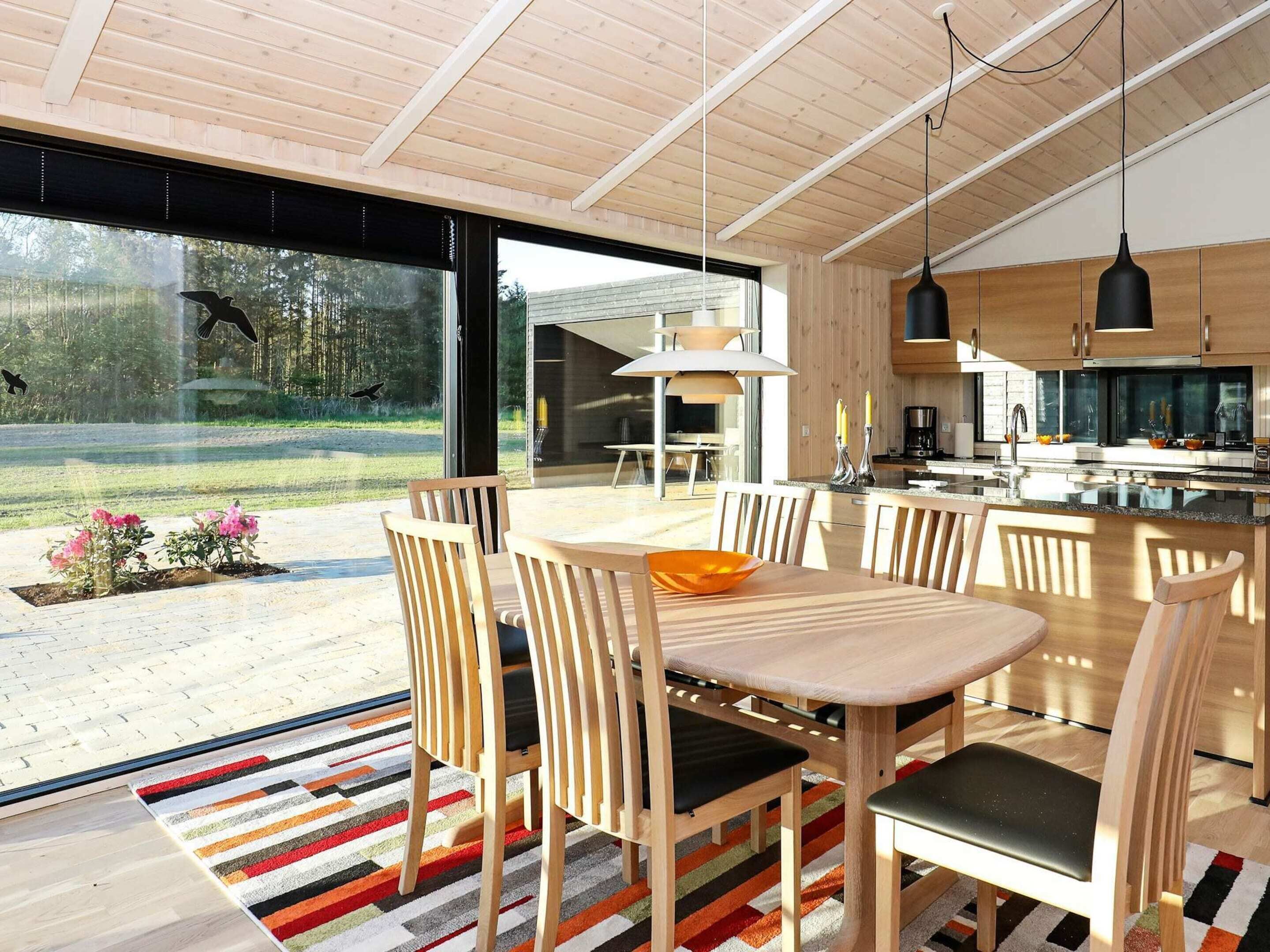 A modern kitchen with wooden cabinets and a large island, a dining area with wooden chairs and a colorful rug, and a large glass door leading to a patio with outdoor seating.