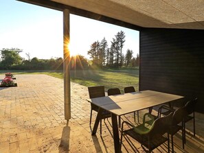 An outdoor patio with a table and chairs, a view of trees and grass, and a building in the background.