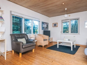 A modern living room with a leather sofa, a wicker chair, a flat-screen TV, and large windows offering a view of trees.