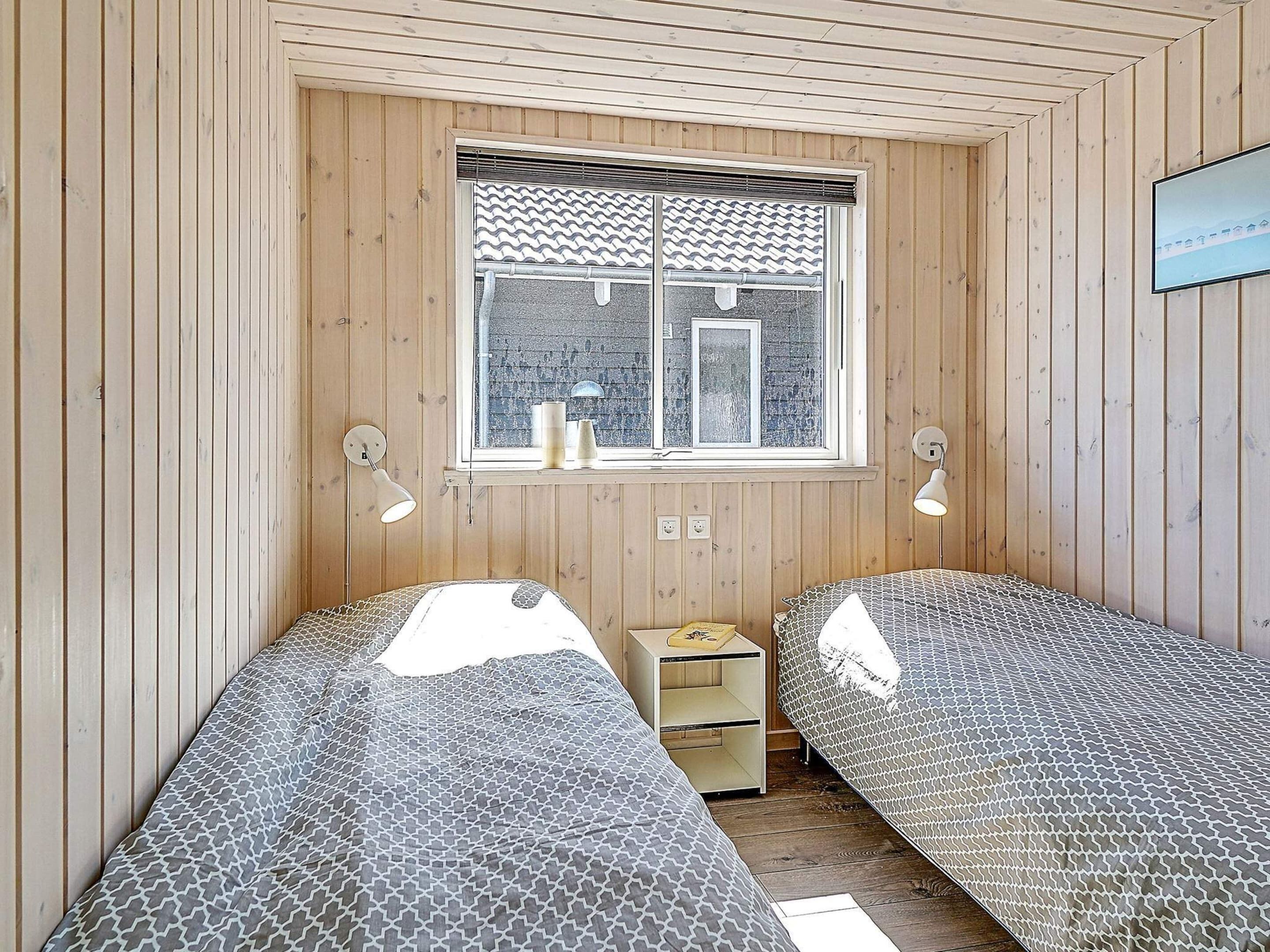 A room with two beds, a window with blinds, a small bedside table, and a wall-mounted lamp.