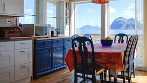 A kitchen with wooden cabinets, a microwave, and a dining table with chairs. There is a view of mountains through large windows. - Holiday Home in DÃ¸nna (Dønna)