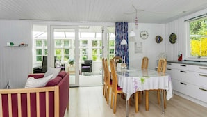 A dining area with a table set for six, surrounded by wooden chairs. There is a sofa with a cushion and a small table with a plant. The room has white walls, a wooden ceiling, and a window with a view of greenery. - Simplistic Holiday Home in Ringkøbing near Sea (Ringkobing)