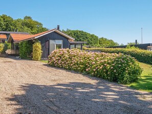 Une allée de gravier mène à une maison avec un toit rouge, entourée d’une végétation luxuriante et d’un grand parterre de fleurs.