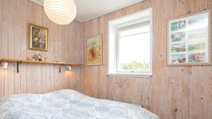 A bedroom with wooden paneling, a bed with a patterned bedspread, a window with a view, and wall-mounted shelves with decorative items. - Spacious Retreat by Dunes - By Traum Ferienwohnungen (Hirtshals)