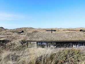 Un bâtiment isolé, d’aspect rustique, doté d’un toit plat et entouré d’herbes sèches et de collines.