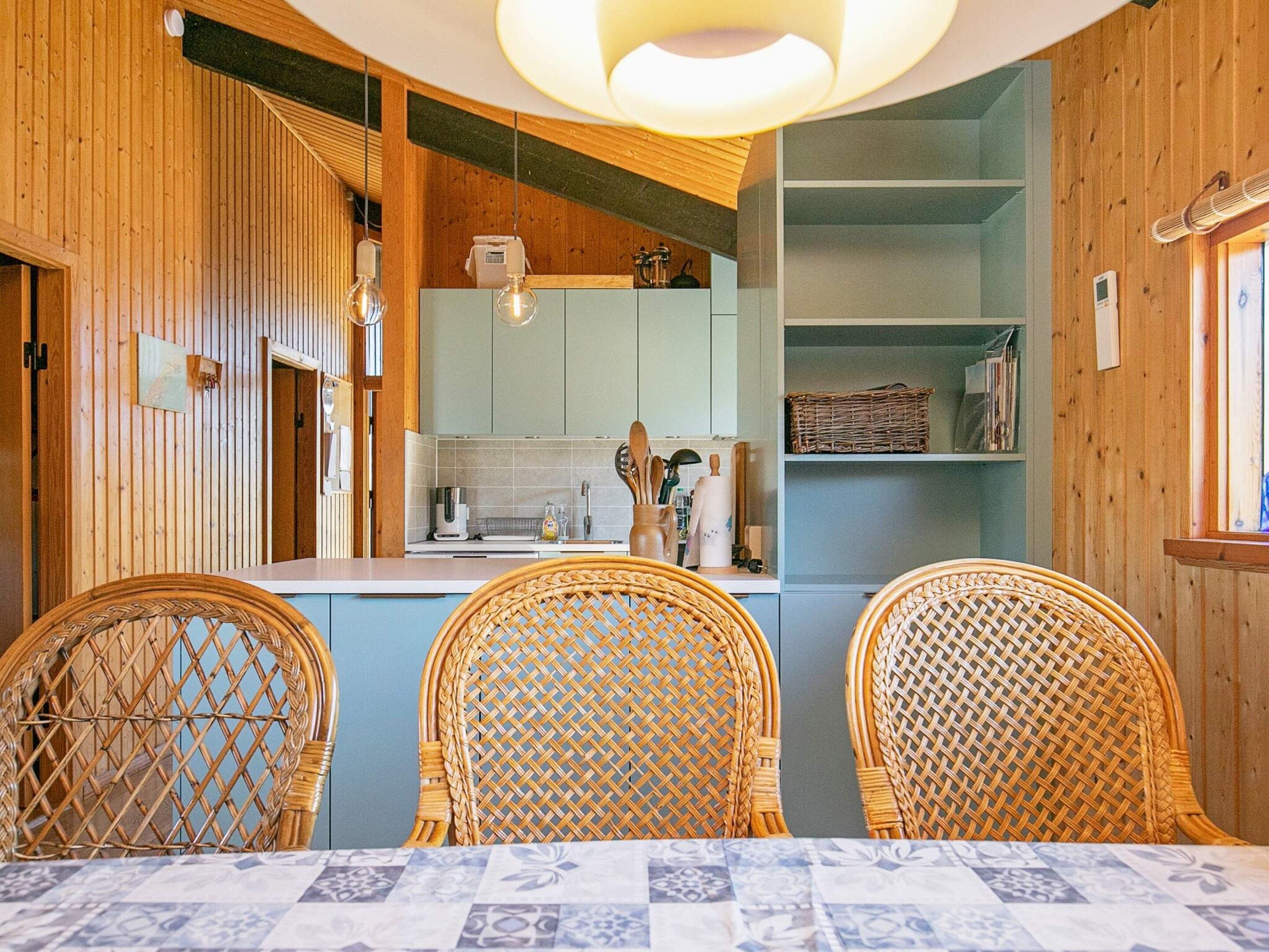 Un coin repas avec des chaises en osier et une table, une cuisine avec des meubles bleu clair et des murs en bois.