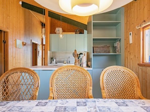 Une cuisine aux murs en bois, avec un plan de travail bleu et des chaises en osier disposées autour d’une table recouverte d’une nappe à motifs.
