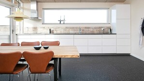 Une cuisine moderne avec des meubles blancs, une grande fenêtre et un coin repas comprenant une table en bois et des chaises marron.