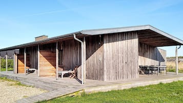 Un edificio moderno con fachada de madera, un patio cubierto con mobiliario exterior y un cielo despejado.