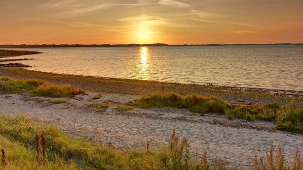 Una playa con arena y césped, una masa de agua y un atardecer.