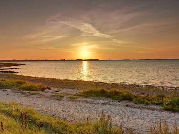 A beach with sand and grass, a body of water, and a sunset.