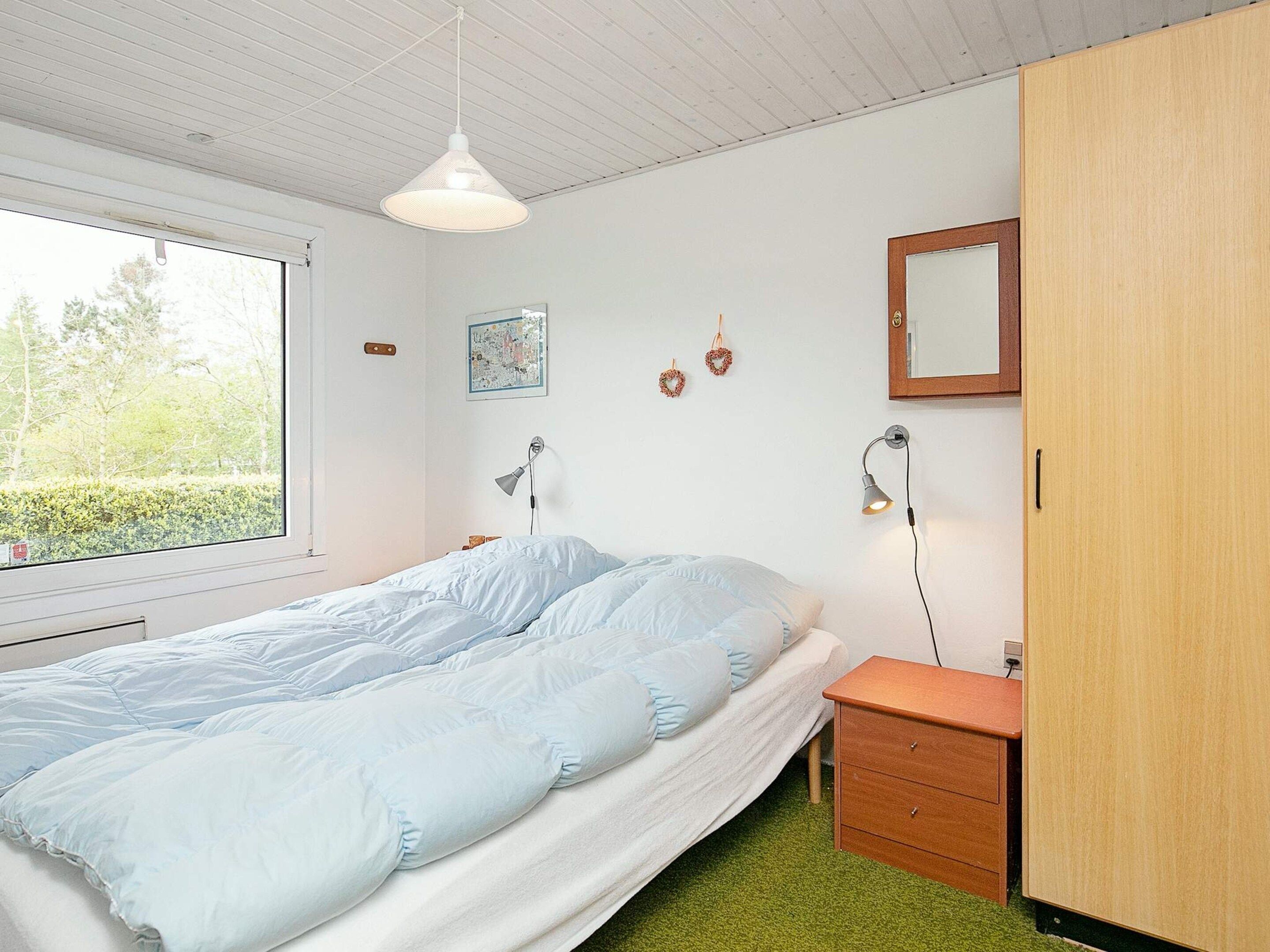 Un dormitorio con cama, mesita de noche, lámpara y ventana con vistas a árboles.