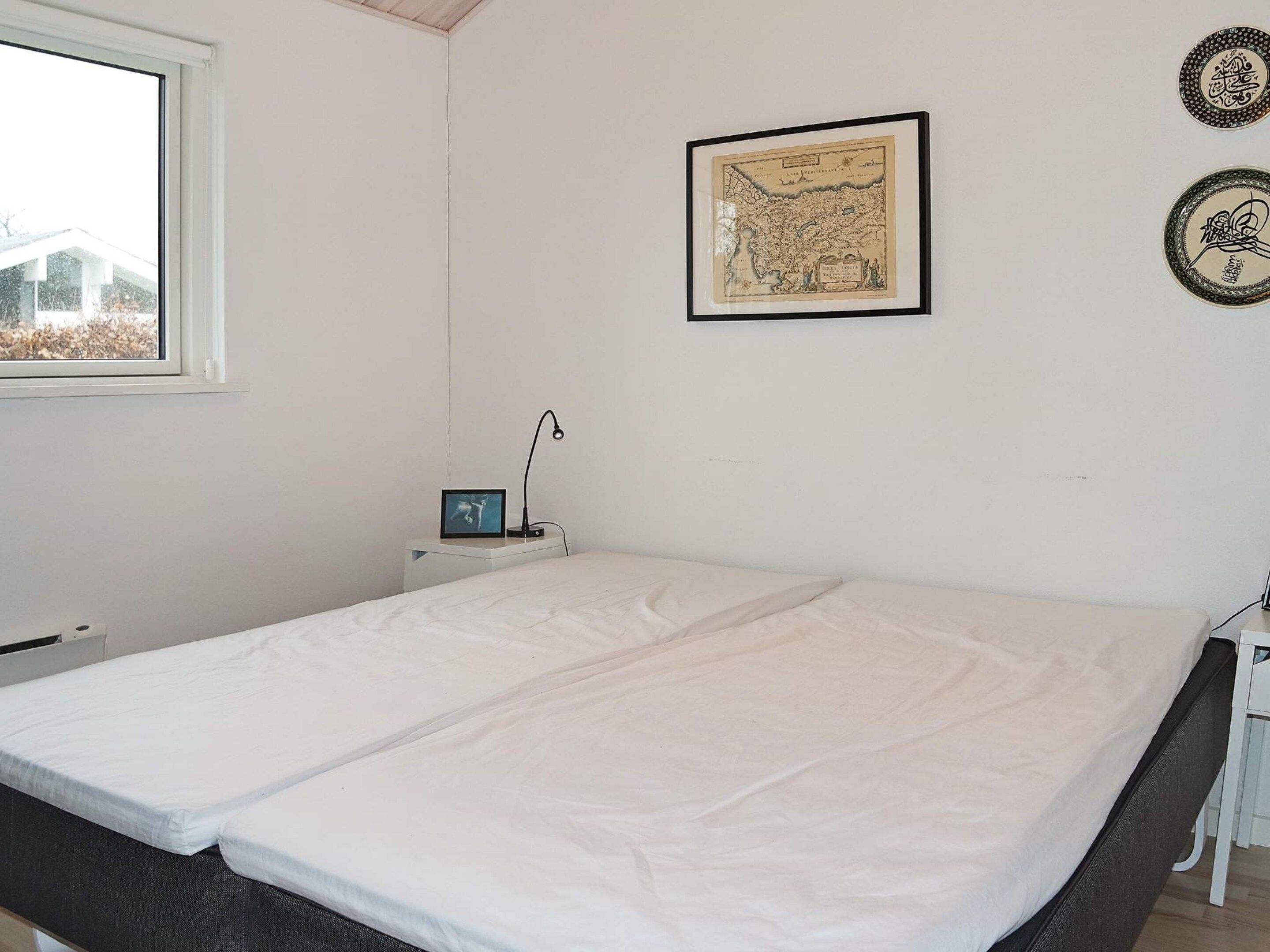 A room with a bed, a desk with a lamp, a framed map on the wall, and a window.