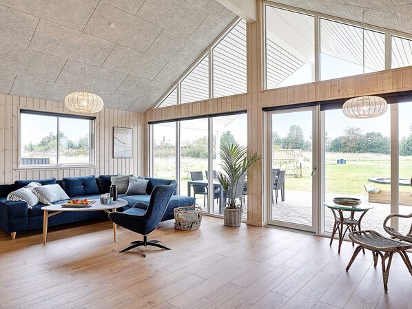 A spacious room with a large sofa, a coffee table, and wicker chairs. There are large windows and a view of the outdoors. - 5 Star Holiday Home in Grenaa (Grenaa)