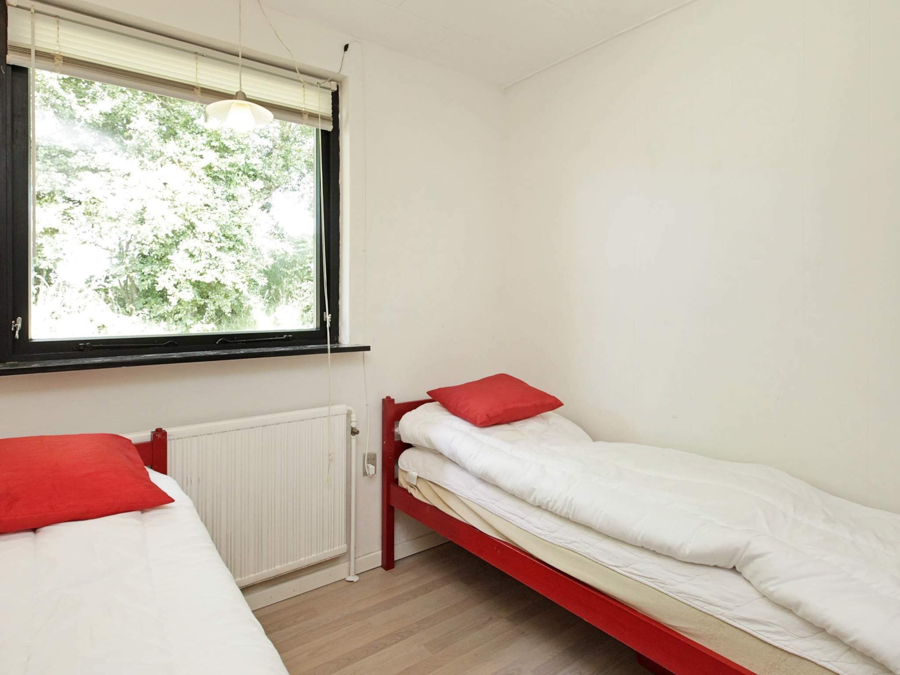 A small, single-room with two beds, a window with blinds, a radiator, and a hanging light fixture.