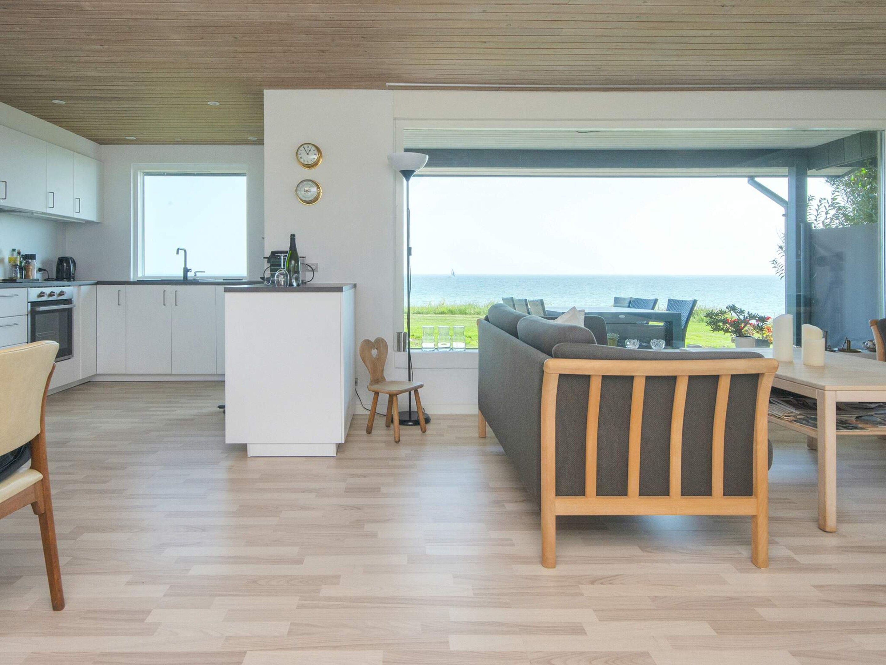 A modern kitchen with white cabinets and a central island, a dining area with a wooden table and chairs, and a large glass door leading to a view of the sea.