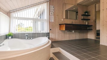 A modern bathroom with a freestanding bathtub, wooden paneling, a large window with blinds, and a dark tiled floor.