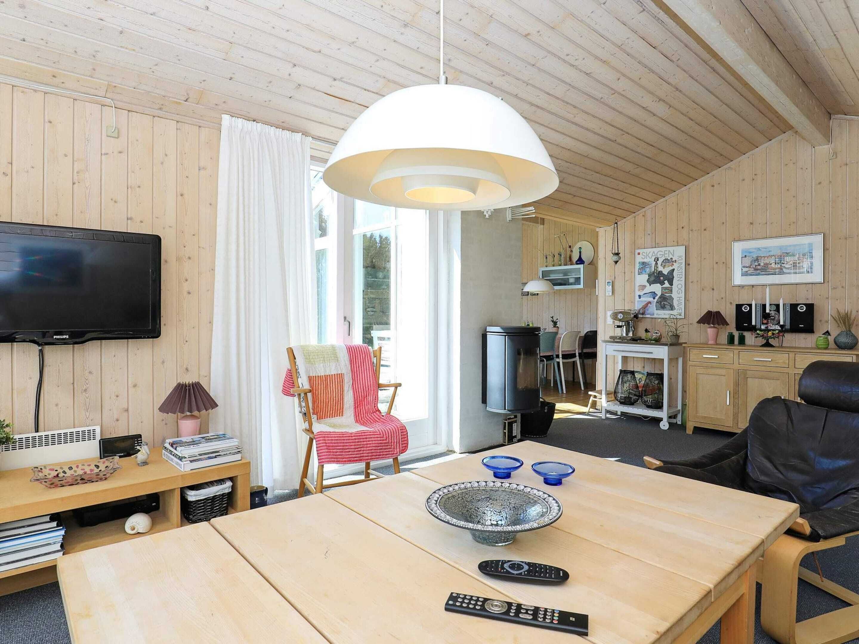 En hyggelig stue med trælofter, et fjernsyn, et spisebord med en skål og fjernbetjeninger, en stol med et stribet tæppe, og et køkkenområde med skabe og en vask.