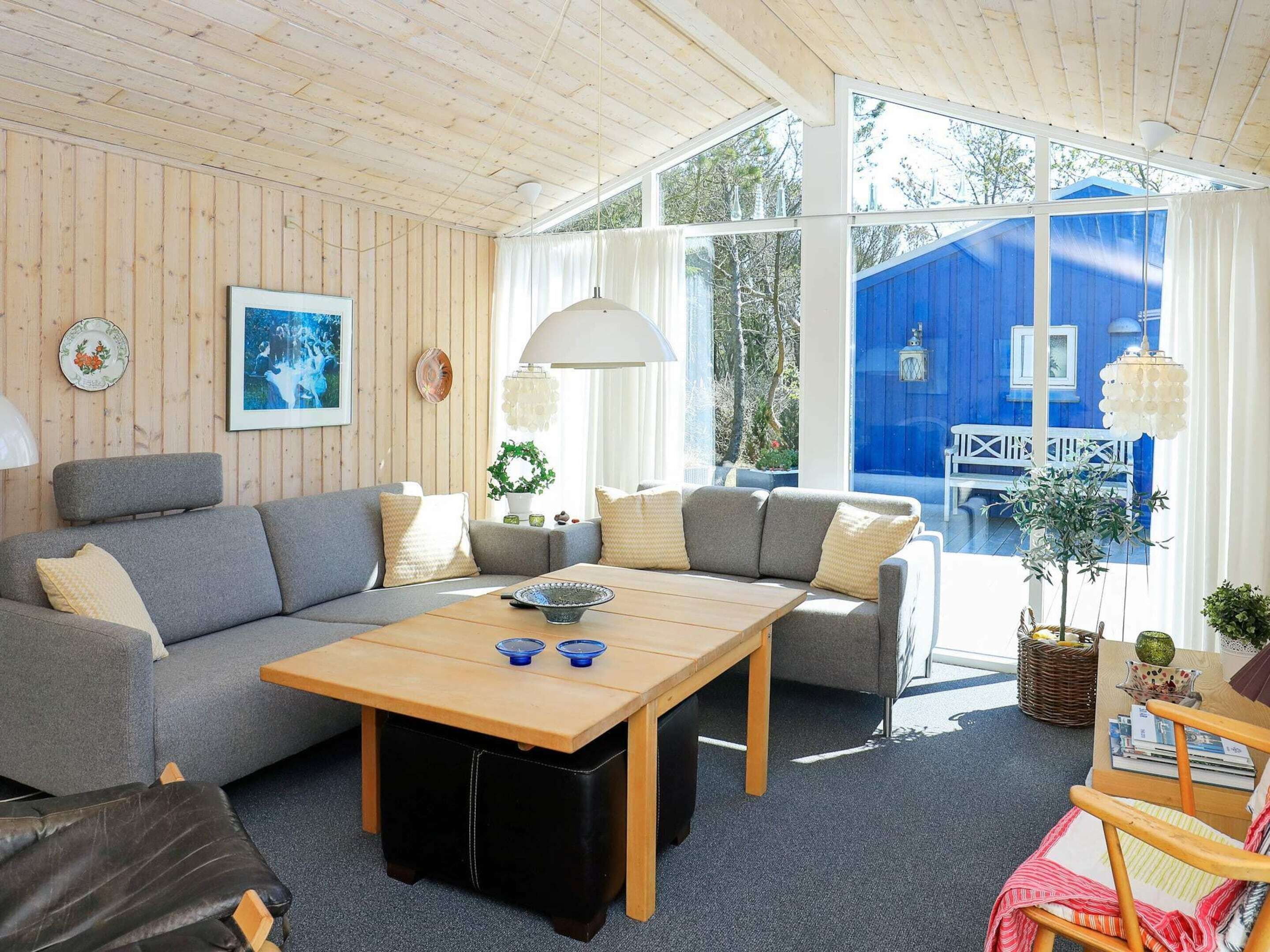 En hyggelig stue med træloft, grå sofaer, et træbord og store vinduer med udsigt til en blå bygning.