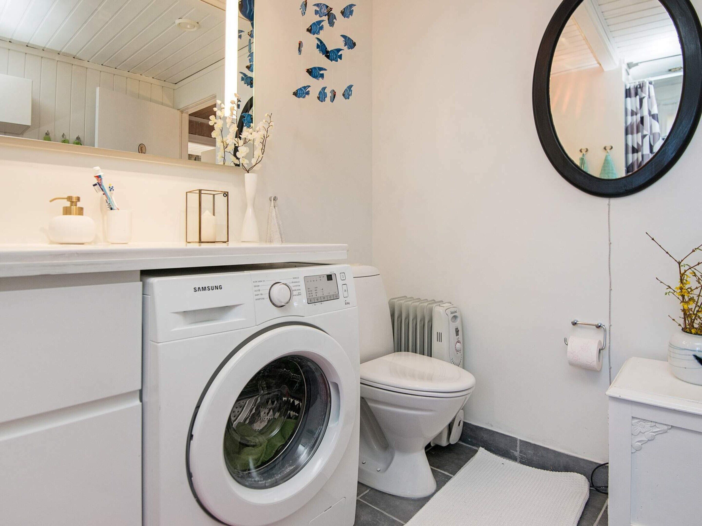 A modern bathroom with a Samsung washing machine, a toilet, a mirror, and a sink with toiletries.