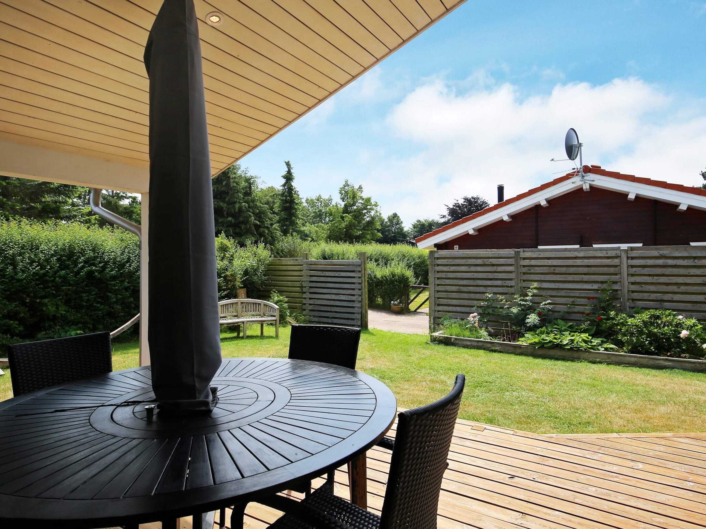 Eine überdachte Terrasse mit rundem Tisch und Stühlen, ein Holzzaun und ein rotes Gebäude im Hintergrund.