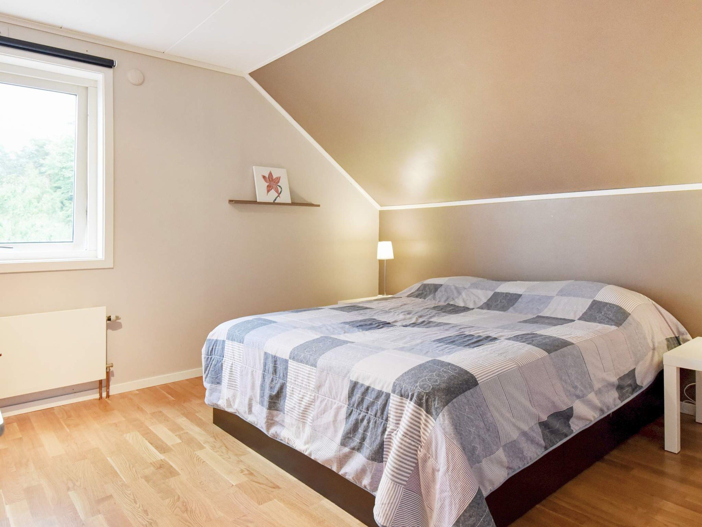 Un dormitorio con cama, mesita de noche, lámpara, radiador, ventana y estante con un cuadro.