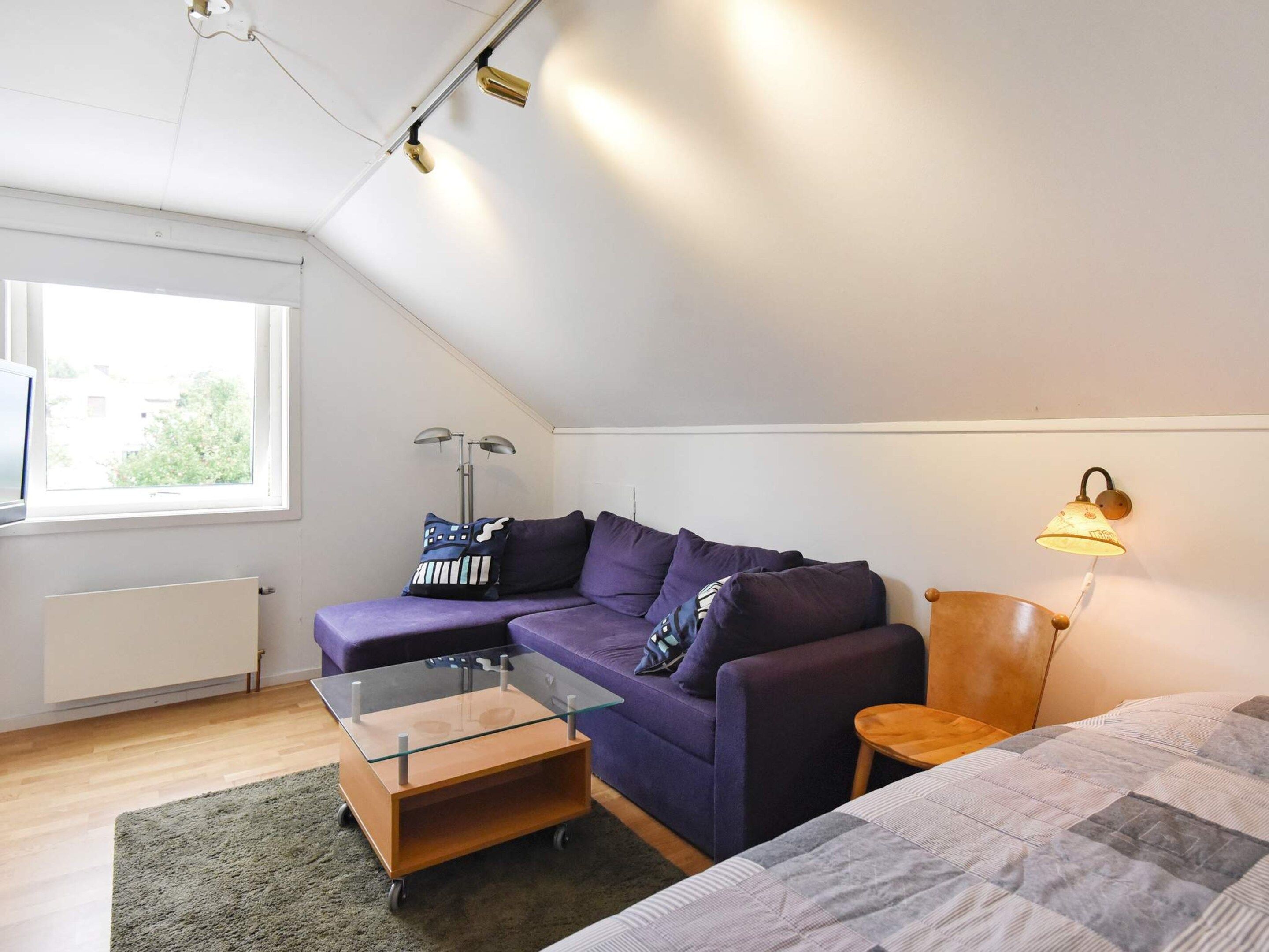 Una habitación moderna y bien iluminada con una mesa de centro de vidrio, un sofá morado con cojines estampados, una mesita de madera y una lámpara de pared.