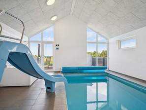An indoor swimming pool with a slide, a blue bench, and a view of the outdoors.