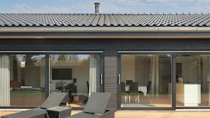 A modern house with a tiled roof, large glass doors, and a wooden deck with two lounge chairs. - 5 Star Holiday Home in Hadsund (Hadsund)