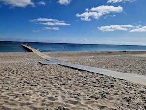 A beach with a wooden walkway leading to the water, a clear sky, and footprints in the sand.