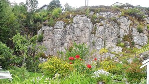 A garden with a bench, a rocky hillside, and a building in the background.