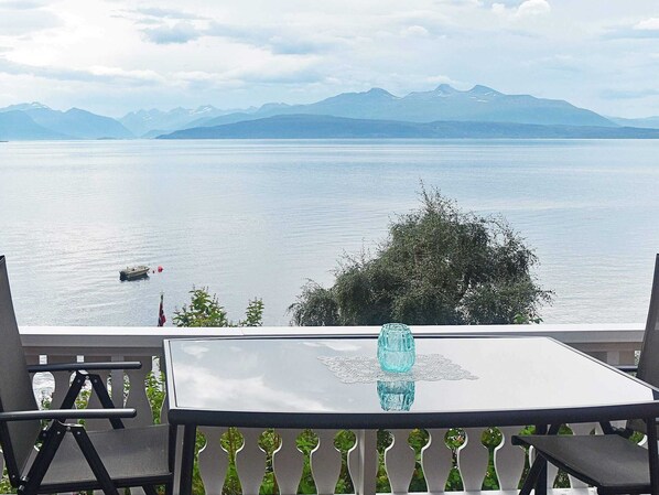 A balcony with a table, chairs, and a view of a lake with mountains in the background.