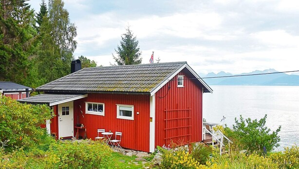 A red wooden cabin with a small porch and two windows, situated by a lake with a rocky path leading to it.