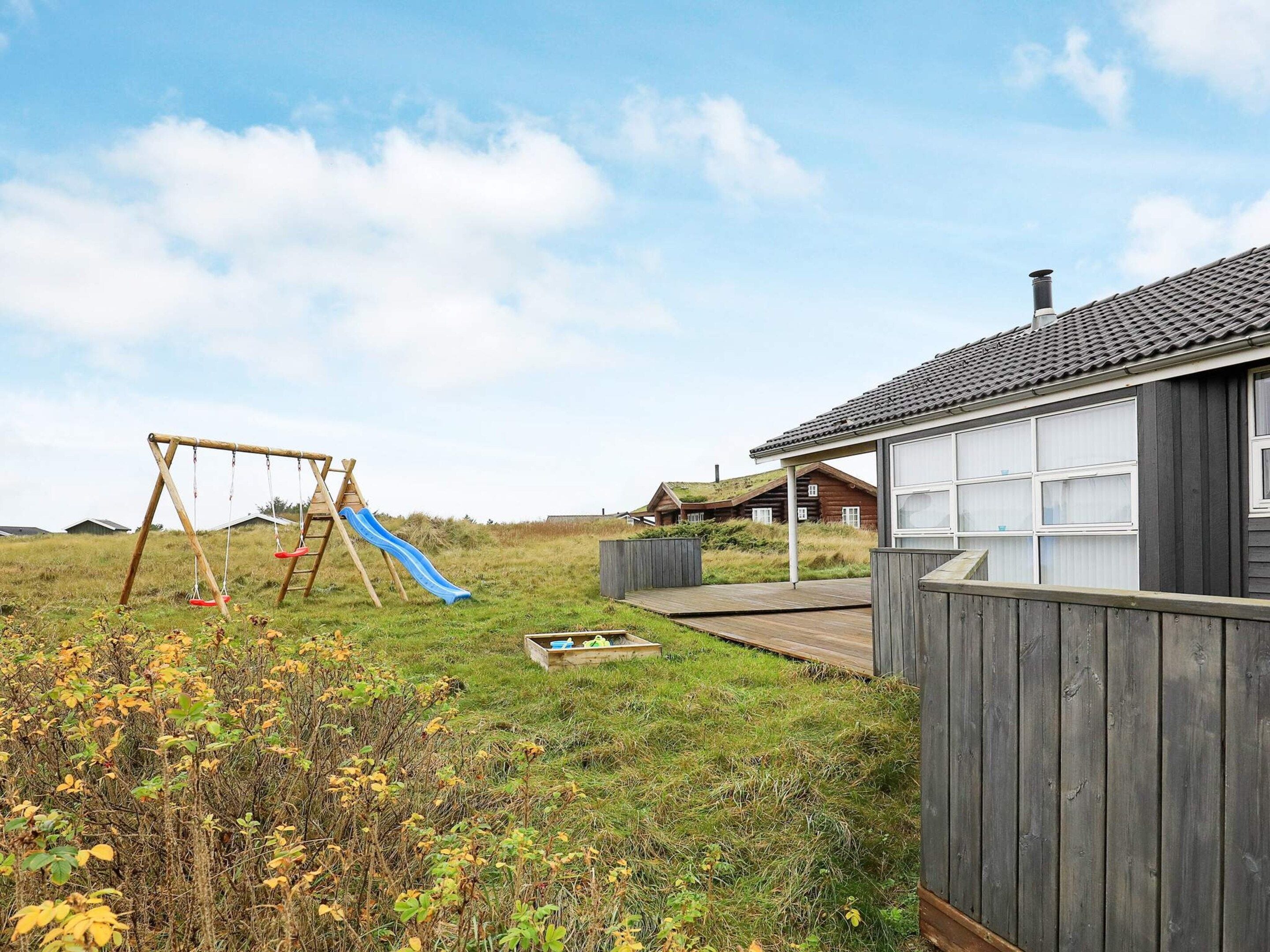 A wooden play structure with a slide and swings, a covered wooden deck, and a house with a dark roof and large windows.
