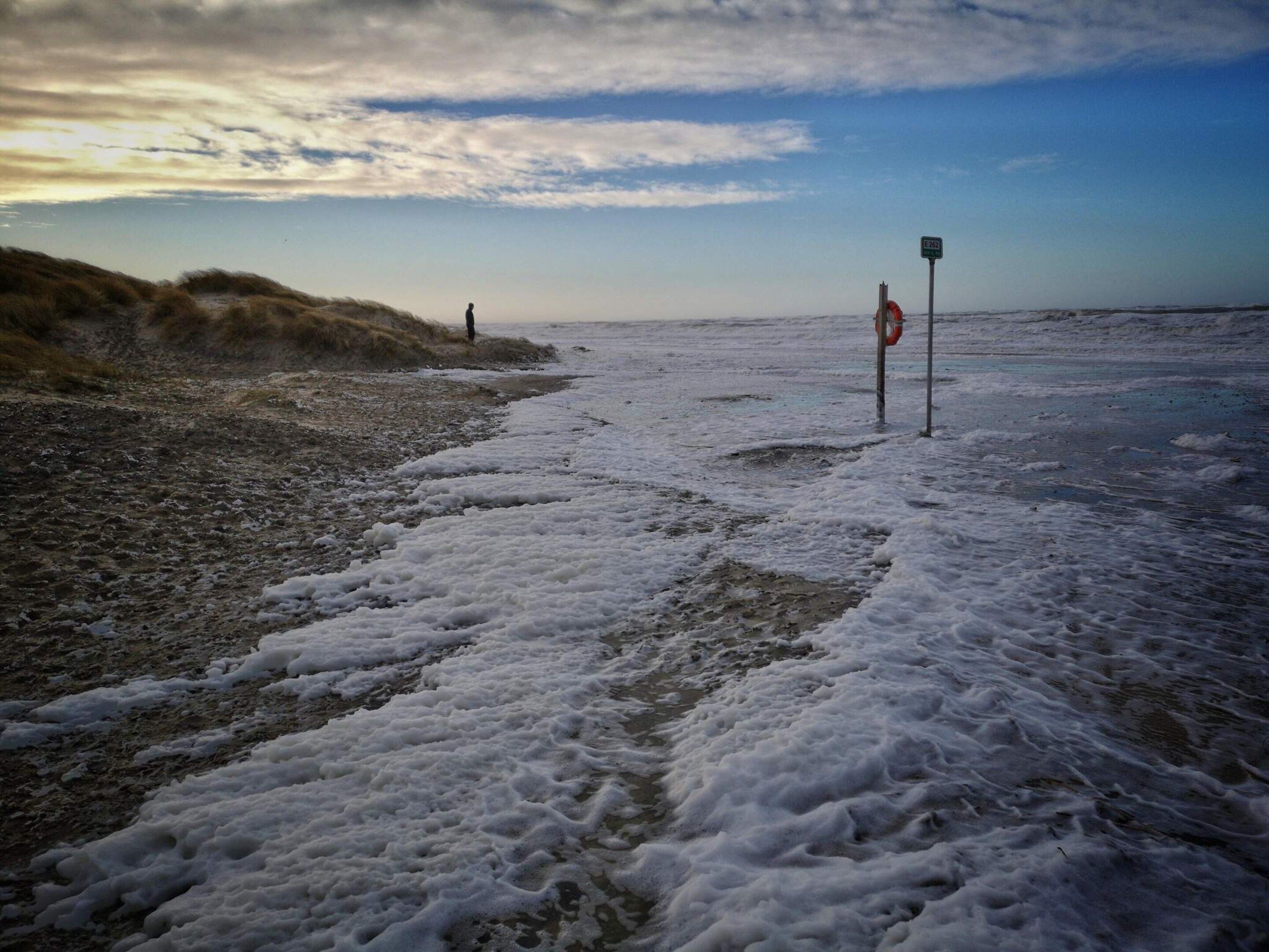 A beach with a lifebuoy and a sign, waves approaching the shore, and a person in the distance.