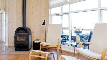 A room with a wood-burning stove, two chairs, a small table, and a view through large windows.