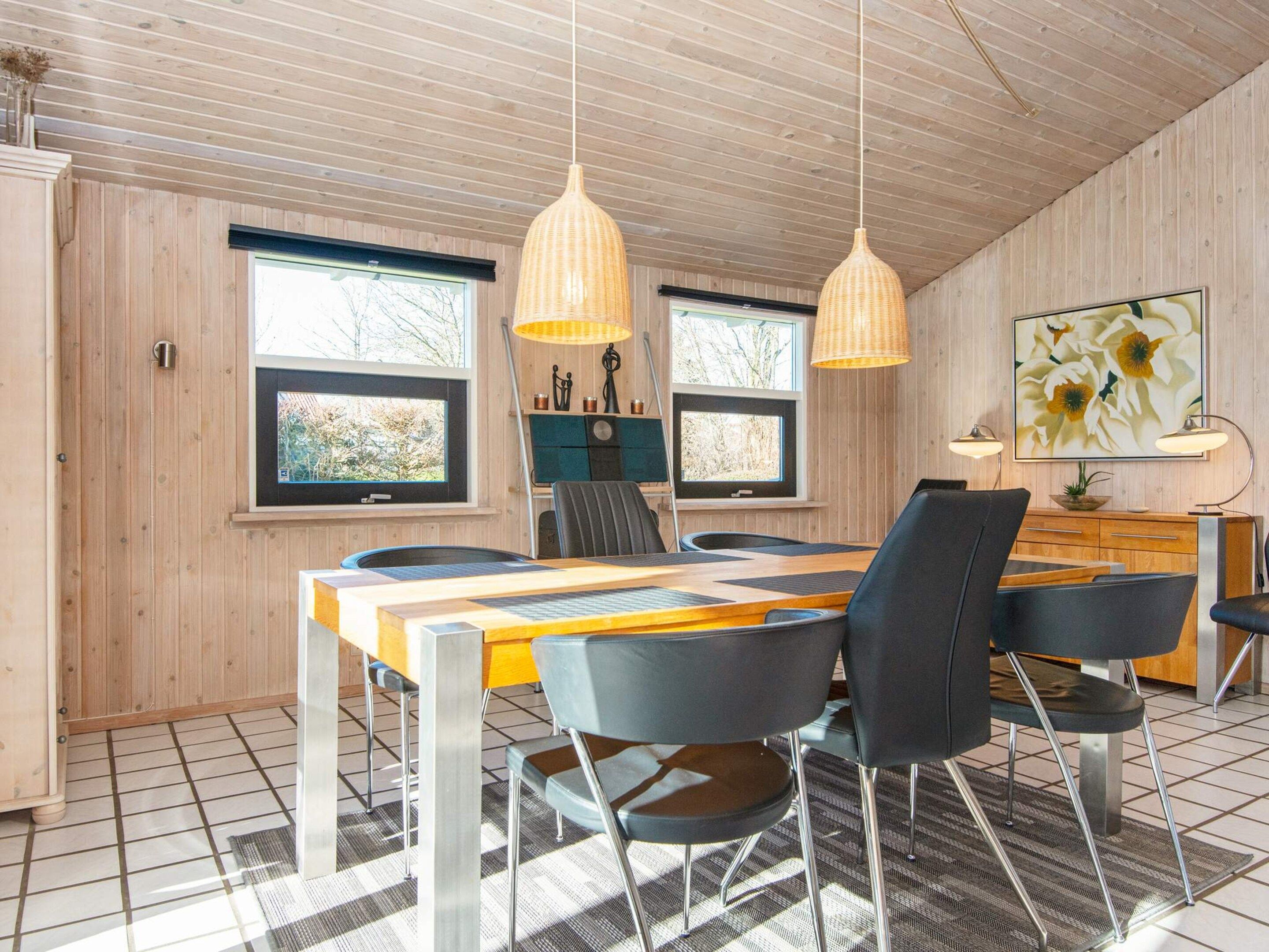 A dining area with a wooden table, chairs, pendant lights, and a view of the outdoors through large windows.