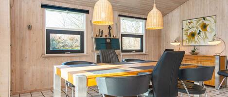 A dining area with a wooden table, chairs, pendant lights, and a view of the outdoors through large windows.