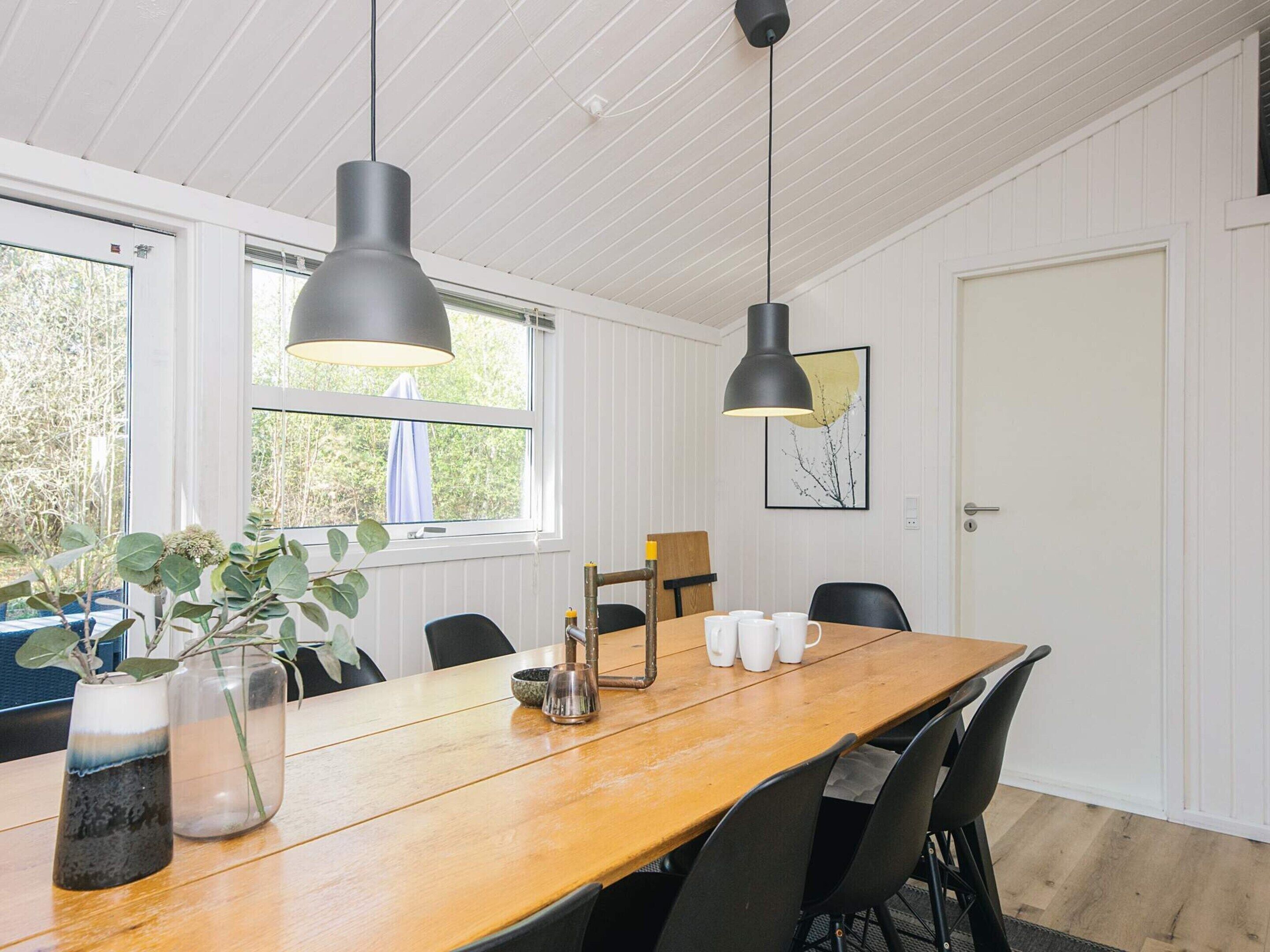 A dining area with a wooden table, black chairs, two pendant lights, a vase with a plant, and a framed picture on the wall.