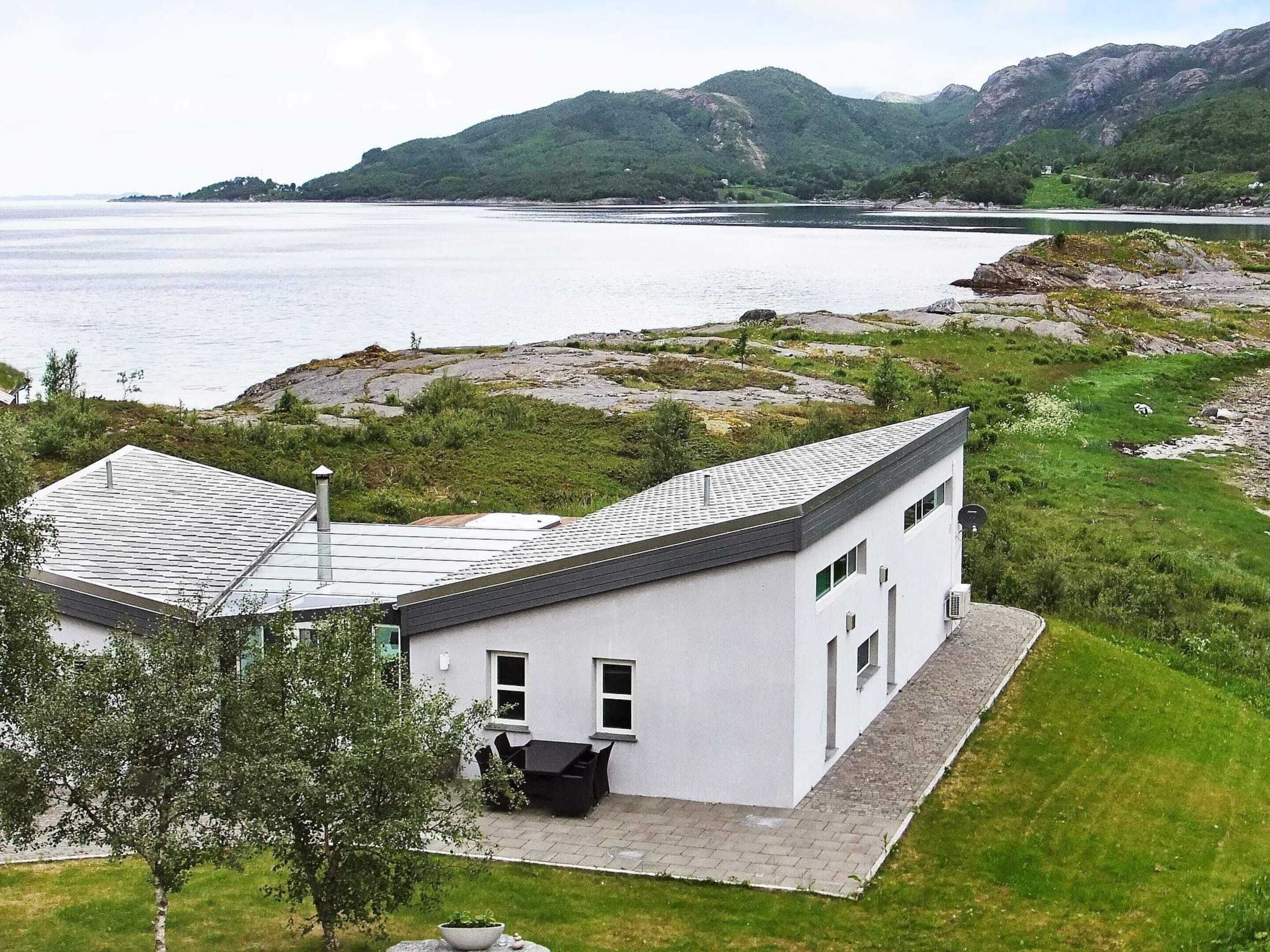 Une maison moderne avec un toit en pente, un petit patio doté de mobilier d’extérieur et une vue sur un lac et des montagnes.