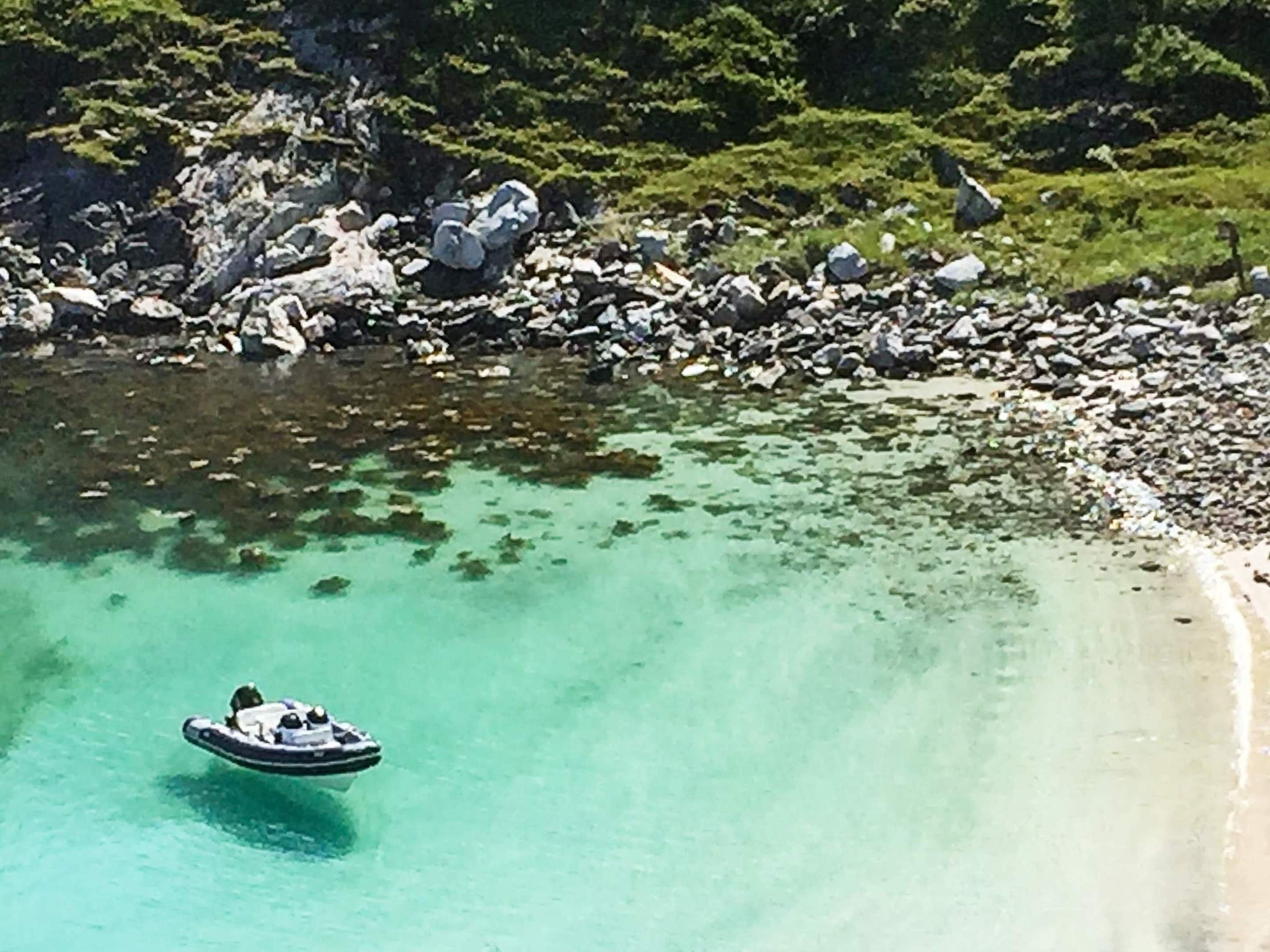 A boat on clear turquoise water near a rocky shoreline with dense greenery in the background.
