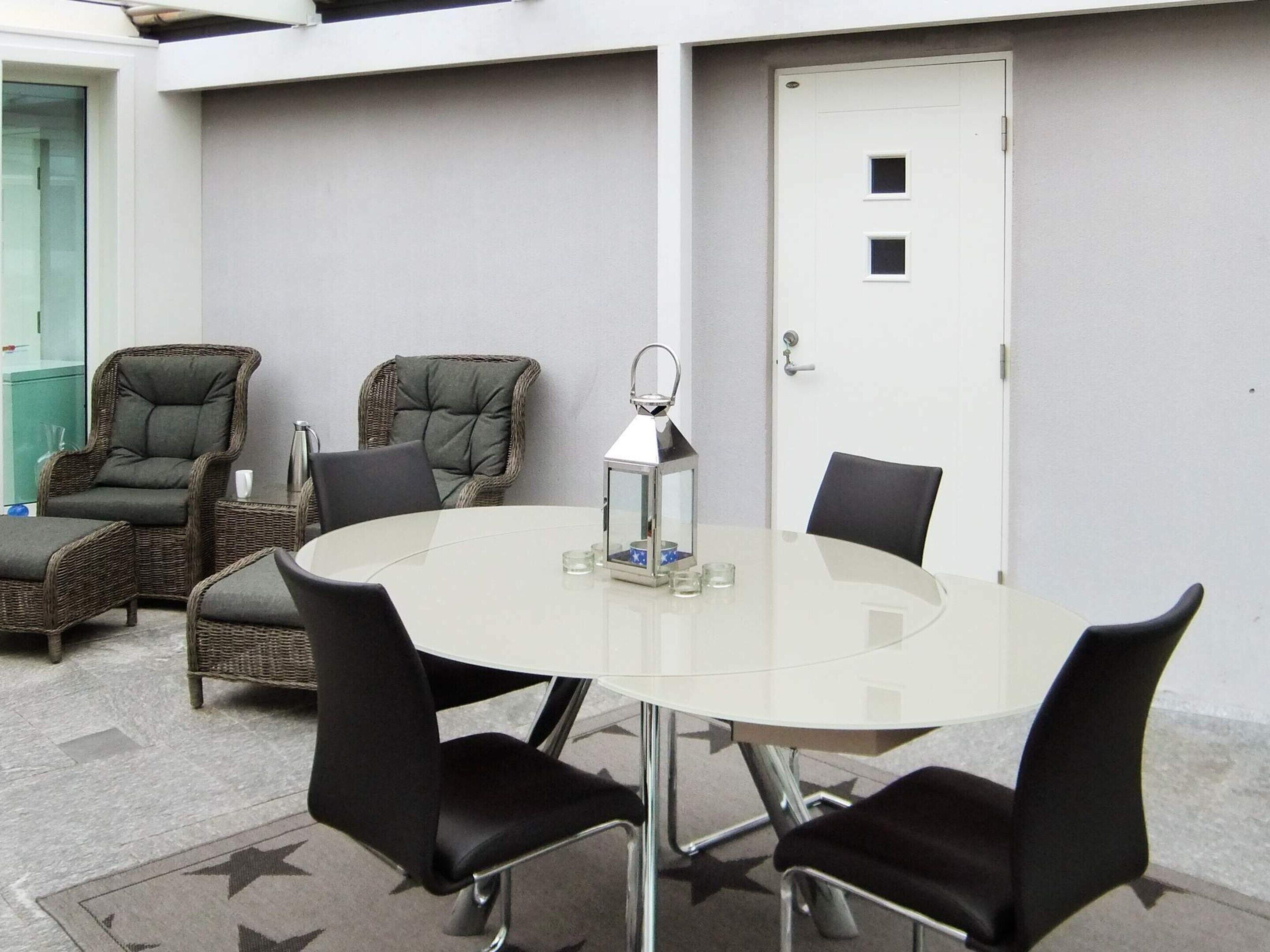 Een vergaderruimte met een glazen tafel, vier zwarte stoelen, twee rieten stoelen, een witte deur en een vloerkleed met stervenspatroon.