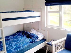 A bunk bed with a blue and white patterned bedspread, a white headboard, and a window with a view of greenery.