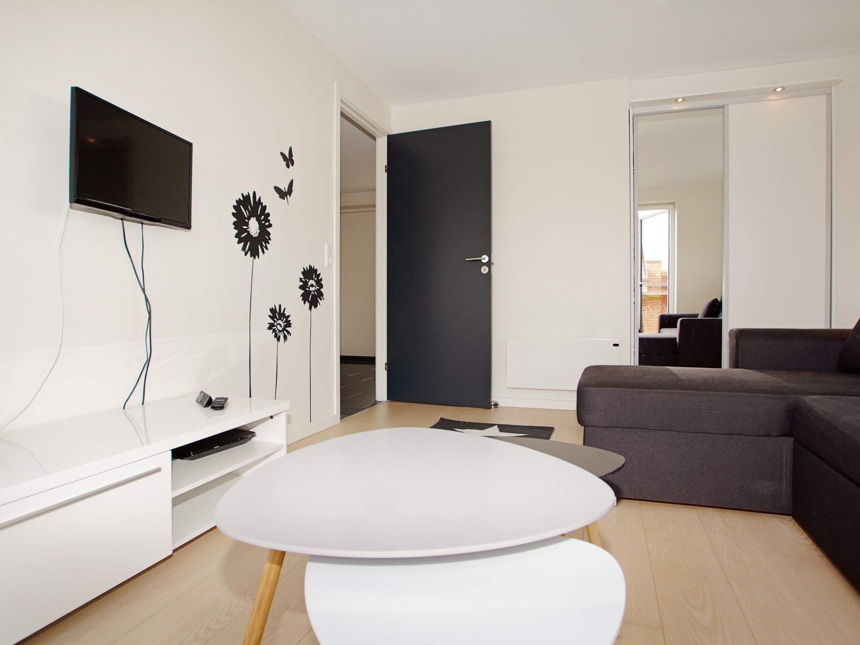 Una sala de estar moderna con un televisor de pantalla plana, una mesa blanca, un sofá oscuro y una puerta que da a otra habitación.