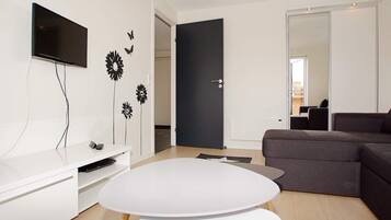 Una sala de estar moderna con un televisor de pantalla plana, una mesa blanca, un sofá oscuro y una puerta que da a otra habitación.