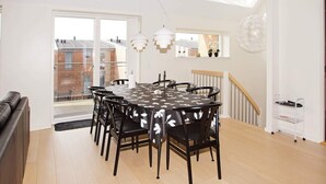 Un coin repas avec une table ronde, des chaises noires et une nappe blanche. On y trouve un escalier avec une rampe, un mur blanc et une fenêtre donnant sur des immeubles.