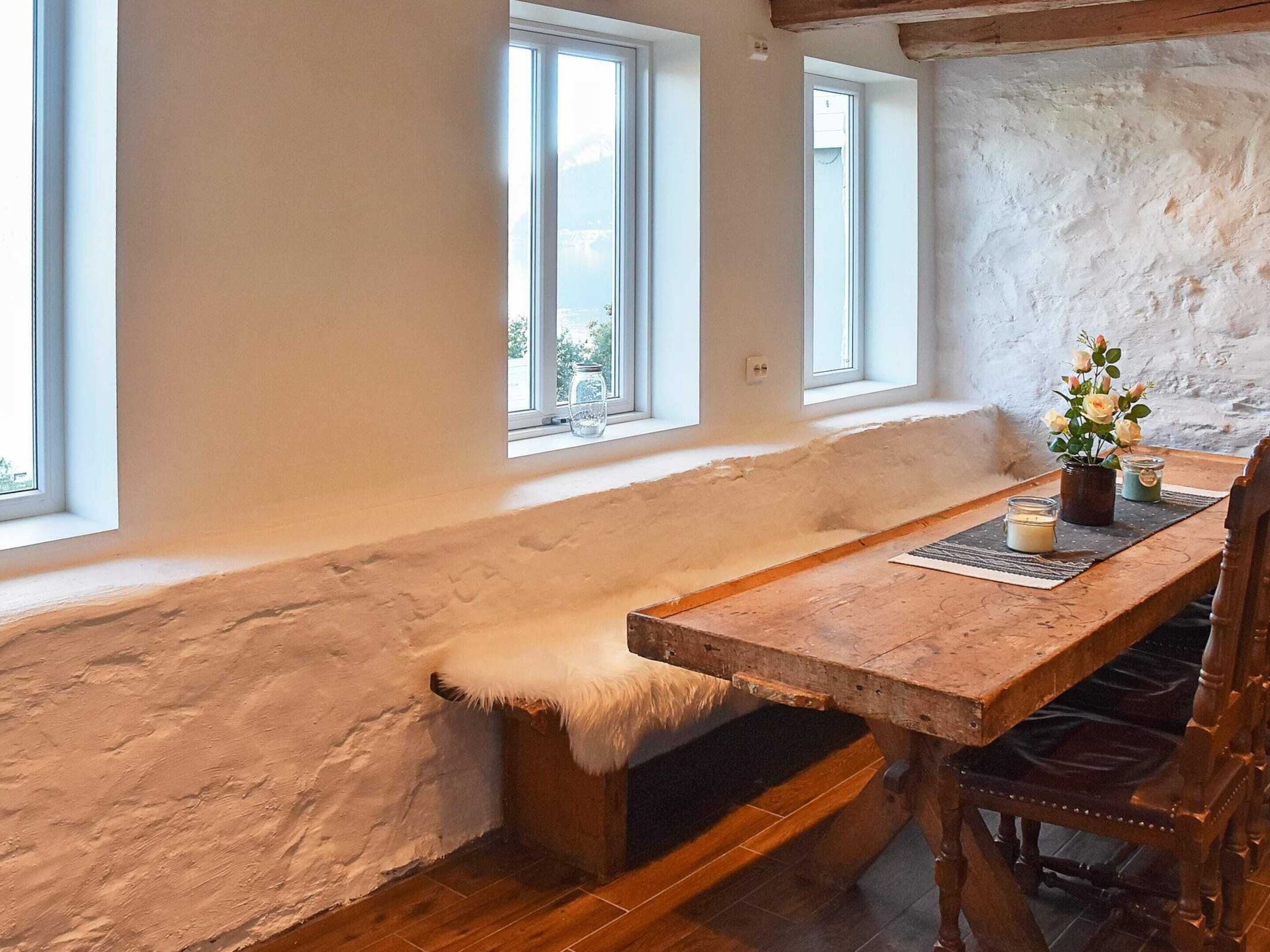 A rustic room with a wooden bench, a table, and a chair. There are three windows providing natural light.