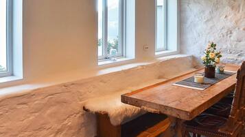 A rustic room with a wooden bench, a table, and a chair. There are three windows providing natural light.